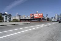 there is an empty highway with white stripes on it with a billboard above it and tall buildings in the distance