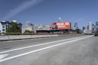 there is an empty highway with white stripes on it with a billboard above it and tall buildings in the distance