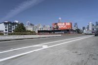 there is an empty highway with white stripes on it with a billboard above it and tall buildings in the distance