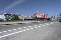 there is an empty highway with white stripes on it with a billboard above it and tall buildings in the distance