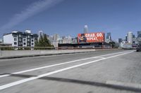 there is an empty highway with white stripes on it with a billboard above it and tall buildings in the distance