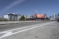there is an empty highway with white stripes on it with a billboard above it and tall buildings in the distance