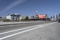 there is an empty highway with white stripes on it with a billboard above it and tall buildings in the distance