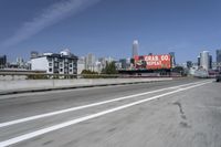 there is an empty highway with white stripes on it with a billboard above it and tall buildings in the distance