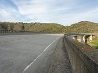 an empty highway near the mountains and bridge at the end of the road as seen from the sidewalk