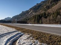 an empty highway with snow and grass on the ground beside a forest with snow capped hills in background