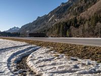 an empty highway with snow and grass on the ground beside a forest with snow capped hills in background