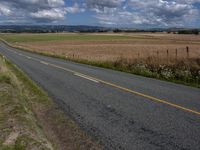 a very empty highway by a wide field with blue skies and clouds above it on the horizon