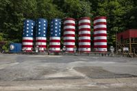 several patriotic cylinders and some trees in the background in an empty lot, with people walking about