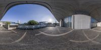 this fisheye lens shows an empty park filled with benches and parked cars under a bridge