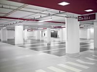 an empty parking garage with multiple columns, red painted ceiling and white floors, light fixtures, and a checkered floor