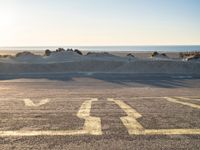 an empty parking lot with sand and water on the side of a beach near the ocean