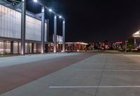 an empty parking lot at night with lights on the building's side and a parking area at the end