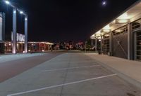 an empty parking lot at night with lights on the building's side and a parking area at the end