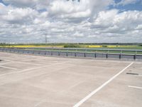 an empty parking lot with a field of green grass and flowers behind the space are blue skies