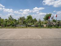 an empty parking lot with benches and flags in the distance, and some weeds in the background