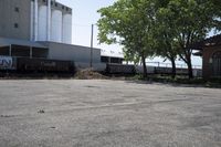 a train is passing by the empty parking lot by a large building and tree branches