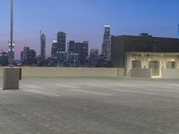 an empty parking lot with a city in the background at dusk while cars are driving down it