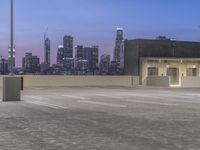 an empty parking lot with a city in the background at dusk while cars are driving down it