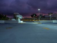 an empty parking lot lit up at night by street lights and purple sky above the buildings