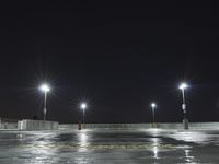 an empty parking lot full of street lights at night time with the ground wet and the roadway reflecting off