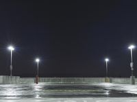 an empty parking lot full of street lights at night time with the ground wet and the roadway reflecting off
