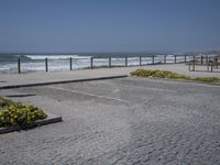 an empty parking lot next to the ocean with several flowers in front of it and a bench overlooking the ocean