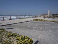 an empty parking lot next to the ocean with several flowers in front of it and a bench overlooking the ocean