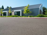 an empty parking lot sits next to a building with a black roof and windows and plants on the side