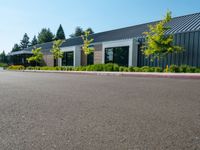 an empty parking lot sits next to a building with a black roof and windows and plants on the side