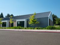 an empty parking lot sits next to a building with a black roof and windows and plants on the side