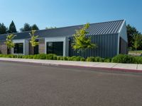 an empty parking lot sits next to a building with a black roof and windows and plants on the side