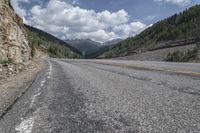 an empty paved mountain road with a rock slope in the foreground and mountains with trees on both sides