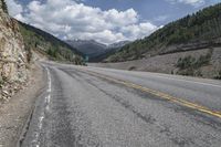 an empty paved mountain road with a rock slope in the foreground and mountains with trees on both sides