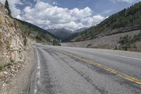 an empty paved mountain road with a rock slope in the foreground and mountains with trees on both sides