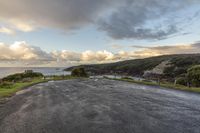 an empty paved road surrounded by hills with ocean in background and clouds in sky over head