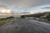 an empty paved road surrounded by hills with ocean in background and clouds in sky over head