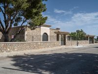 an empty paved street with stone fence and trees lining both sides of the road, near a villa