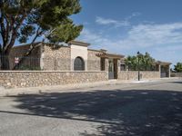 an empty paved street with stone fence and trees lining both sides of the road, near a villa