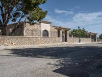 an empty paved street with stone fence and trees lining both sides of the road, near a villa