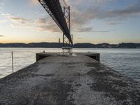 an empty pier and some very pretty water under a big bridge and buildings and a cloudy sky