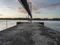 an empty pier and some very pretty water under a big bridge and buildings and a cloudy sky