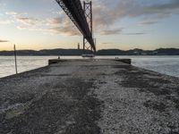 an empty pier and some very pretty water under a big bridge and buildings and a cloudy sky