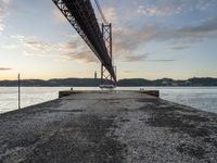 an empty pier and some very pretty water under a big bridge and buildings and a cloudy sky