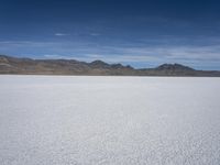 white snow is spread across the flat surface of an empty plain, with mountains in background