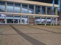 an empty plaza with large windows and people walking along it outside of it at night