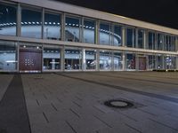 an empty plaza with large windows and people walking along it outside of it at night