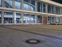 an empty plaza with large windows and people walking along it outside of it at night