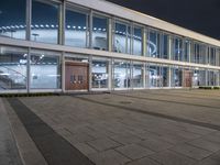 an empty plaza with large windows and people walking along it outside of it at night