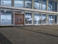 an empty plaza with large windows and people walking along it outside of it at night